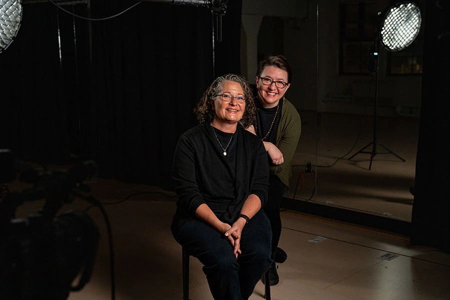 Portrait of Alexis Black sitting and Tina M. Newhauser standing behind her in a black box theatre.