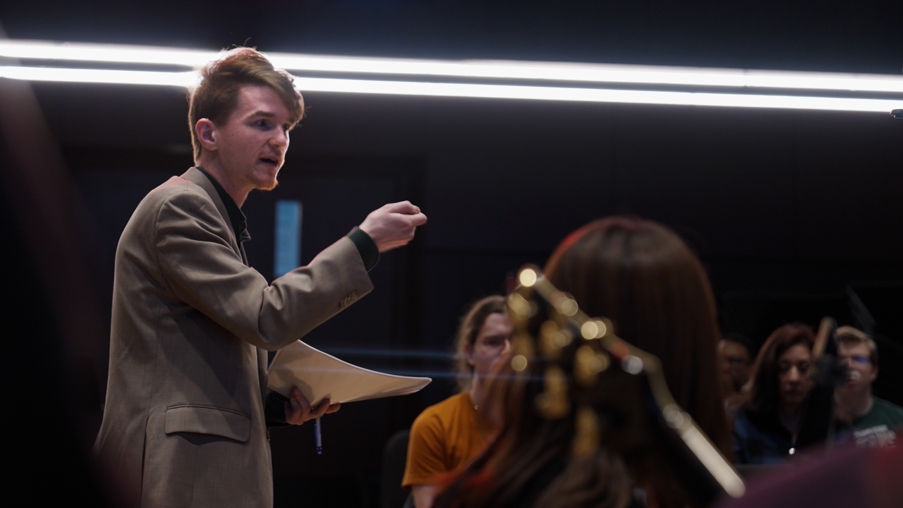 DMA student Tyler Mazone, wearing a grey suit jacket, raises his hand to conduct student musicians.