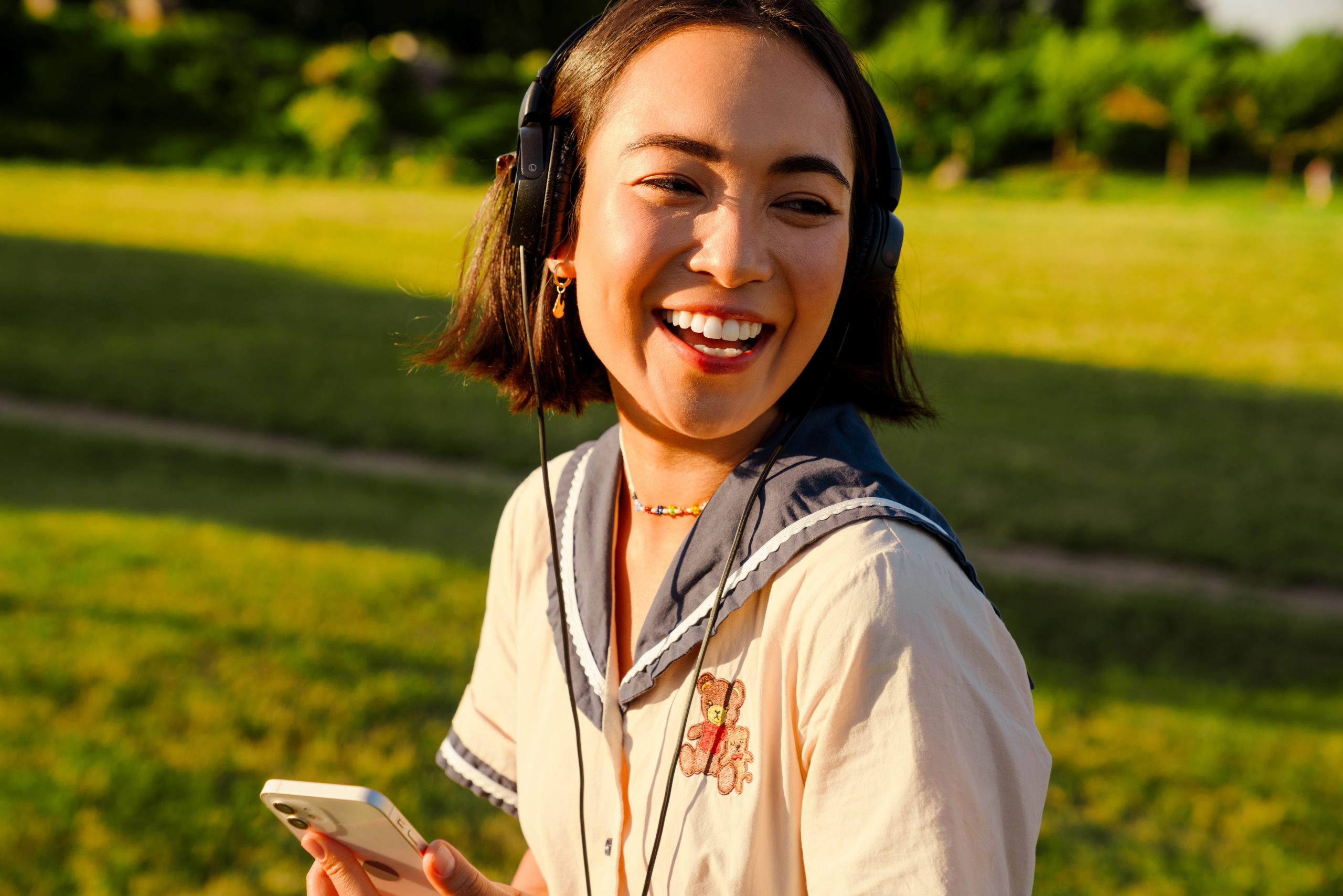 Cheerful young asian girl in headphones using mobile phone during walk outside