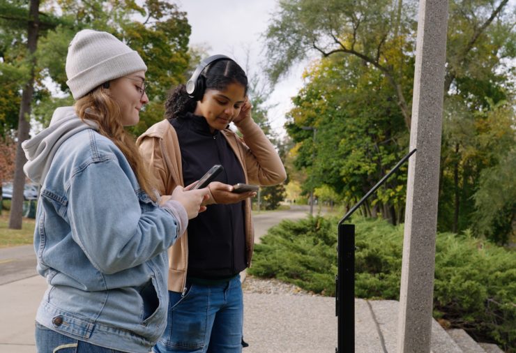 Two young women stand on the Red Cedar River Trail engaged with their smartphones. One woman, wearing a light blue denim jacket and a white beanie, looks at her phone with a smile. The other woman, in a brown jacket and black shirt, adjusts her headphones while focusing on her device.