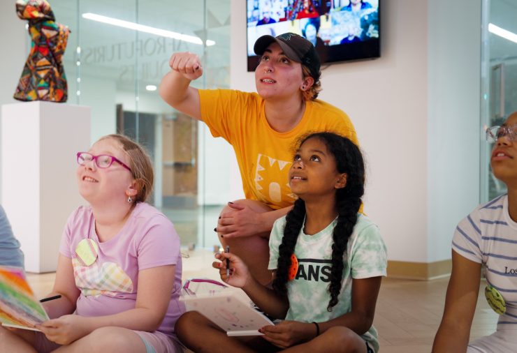 Emily Sylman in a yellow shirt engages a group of young students sitting with sketchbooks, attentively observing an unseen artwork in an art gallery.