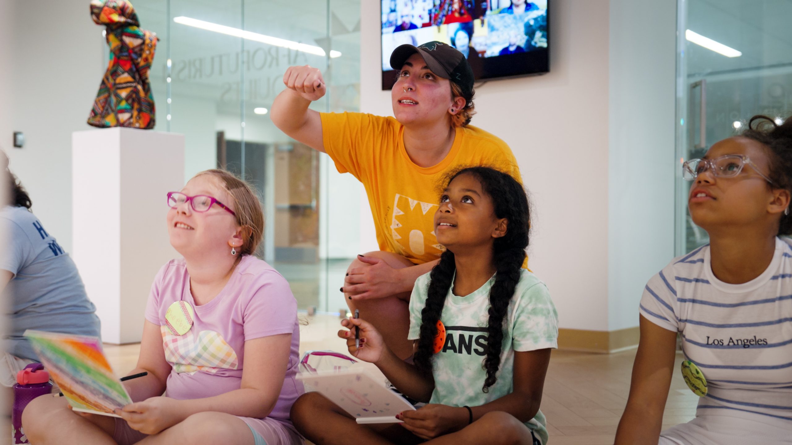 Emily Sylman in a yellow shirt engages a group of young students sitting with sketchbooks, attentively observing an unseen artwork in an art gallery.