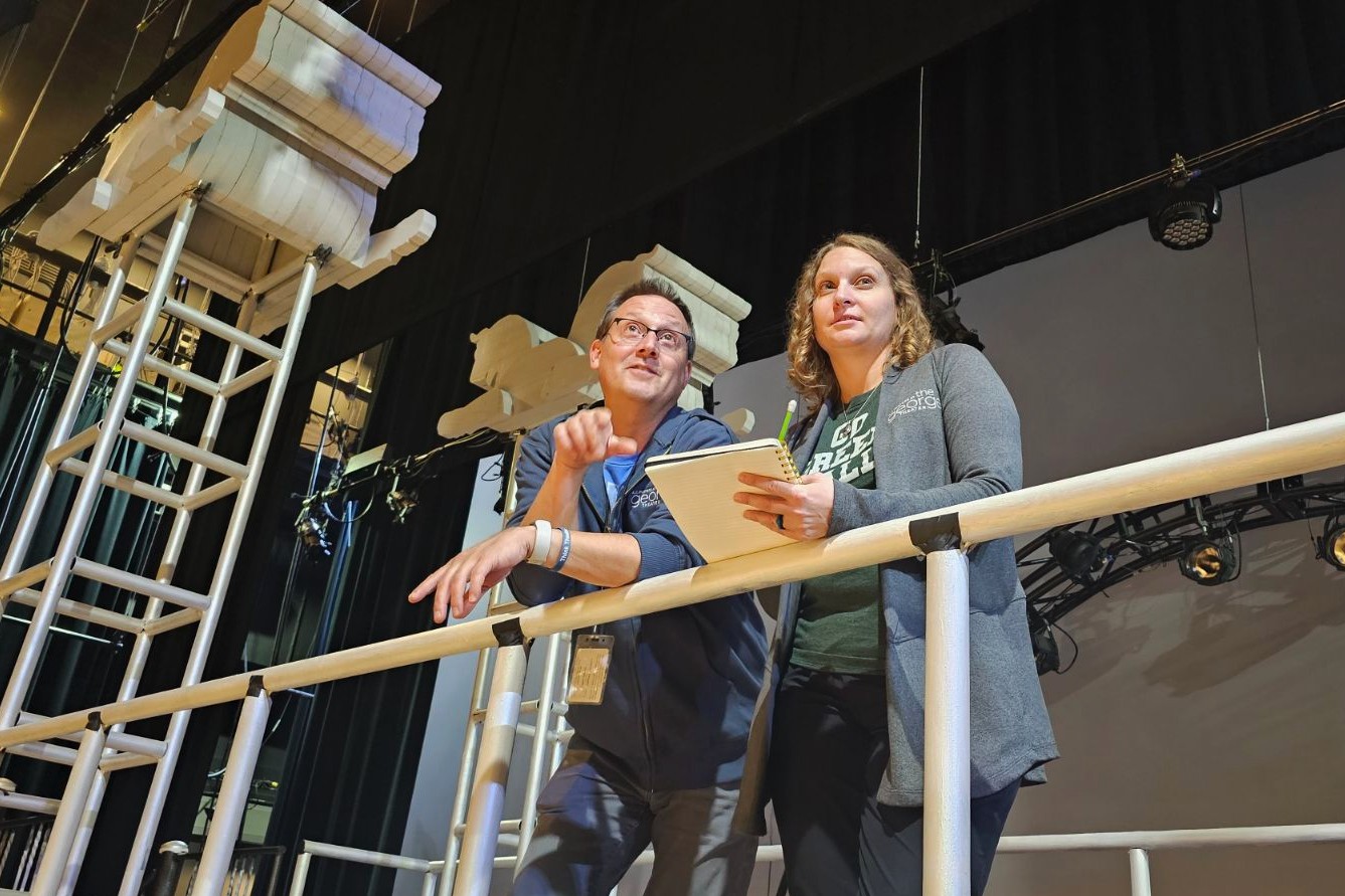 Two people standing on a set platform with stage light overhead. The on the left gestures while the woman on the right holds a notepad. The set features large classical column pieces and ladders in the background.