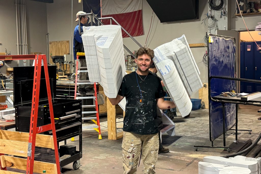 A smiling man in a paint-splattered shirt holds two large, foam architectural pieces resembling classical columns in each arm. The workshop behind him has tools, ladders, and equipment scattered around, with another person working in the background.