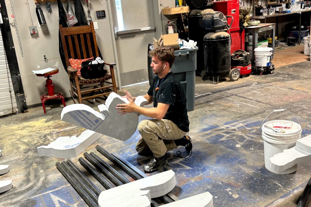 A man kneels on the floor of a workshop, working on a large foam piece shaped like part of a classical column. Surrounding him are additional pieces, buckets of paint, and various tools.