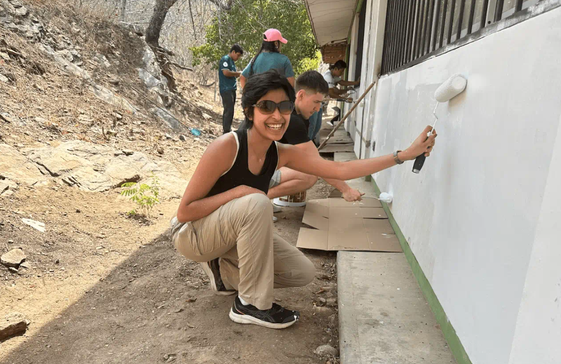 Person smiling while painting the exterior wall of a building white using a roller brush. They are outdoors on a sunny day, with other people in the background also painting and working.