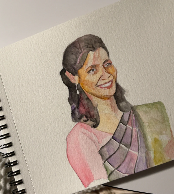 A watercolor painting of a smiling woman wearing a pink and patterned saree. The artwork is created on texted paper and displayed in a spiral sketchbook.