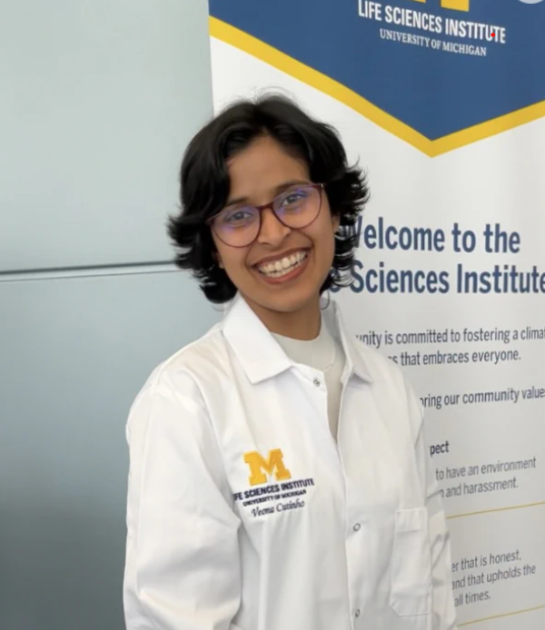 Veona smiling in a lab coat in front of a University of Michigan Life Sciences Institute banner.