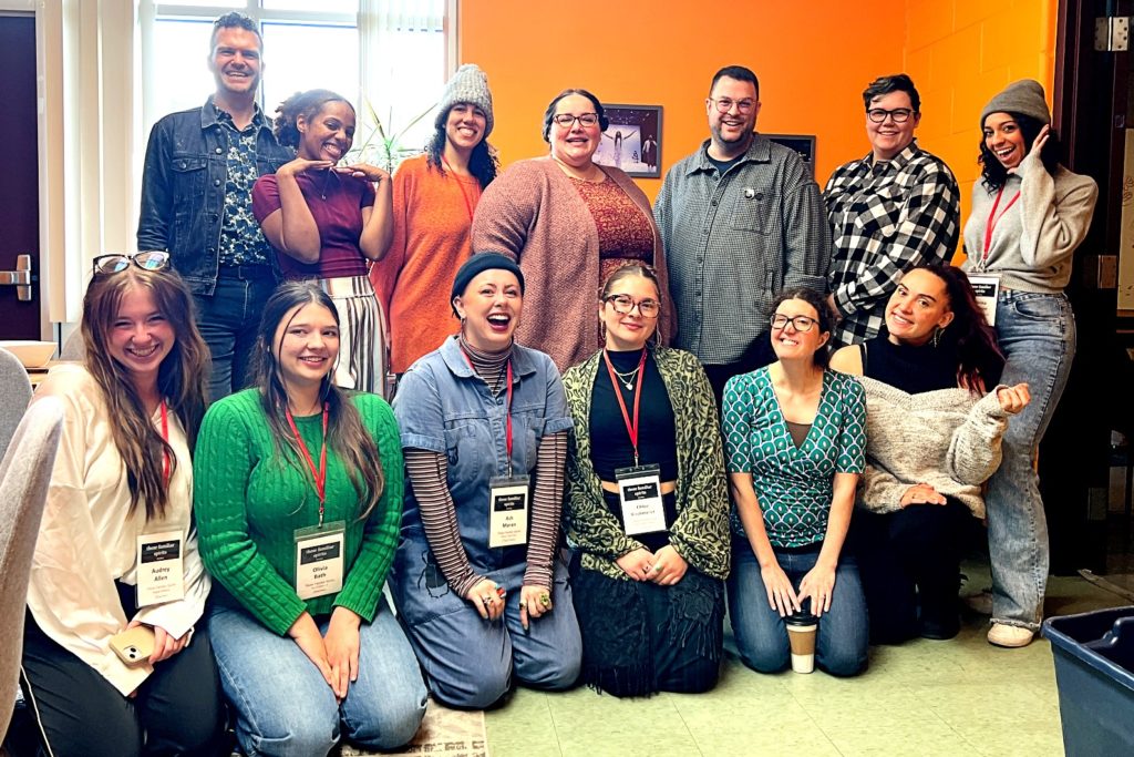 A group of 13 people posing for a photo indoors, smiling and playful. The group is diverse in age and style, with some seated and others standing. The background is a bright orange wall, a window, and framed artwork.