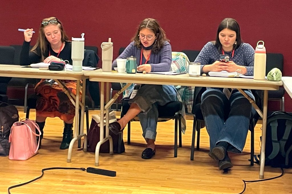 A picture of three individuals seated at a long table with papers, notebooks, and beverages in front of them. From left to right: one person is writing, another is speaking, and the third is focused on their phone. The room has red walls and a polished wood floor.