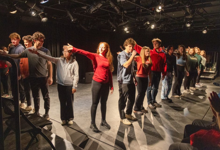 A group of performers are lined up in a V-shape on a black theatre stage.