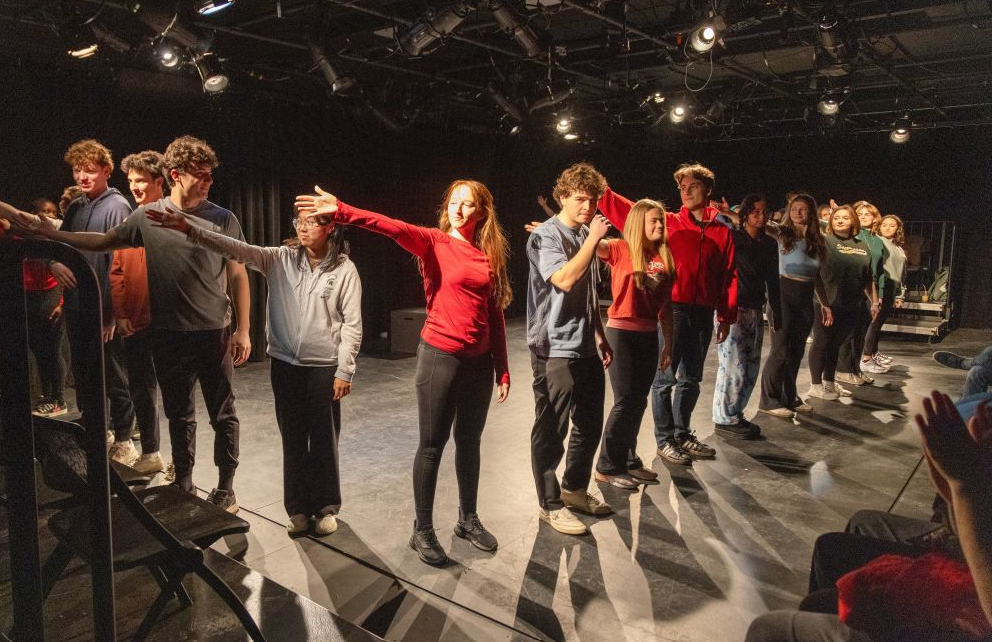 A group of performers are lined up in a V-shape on a black theatre stage.