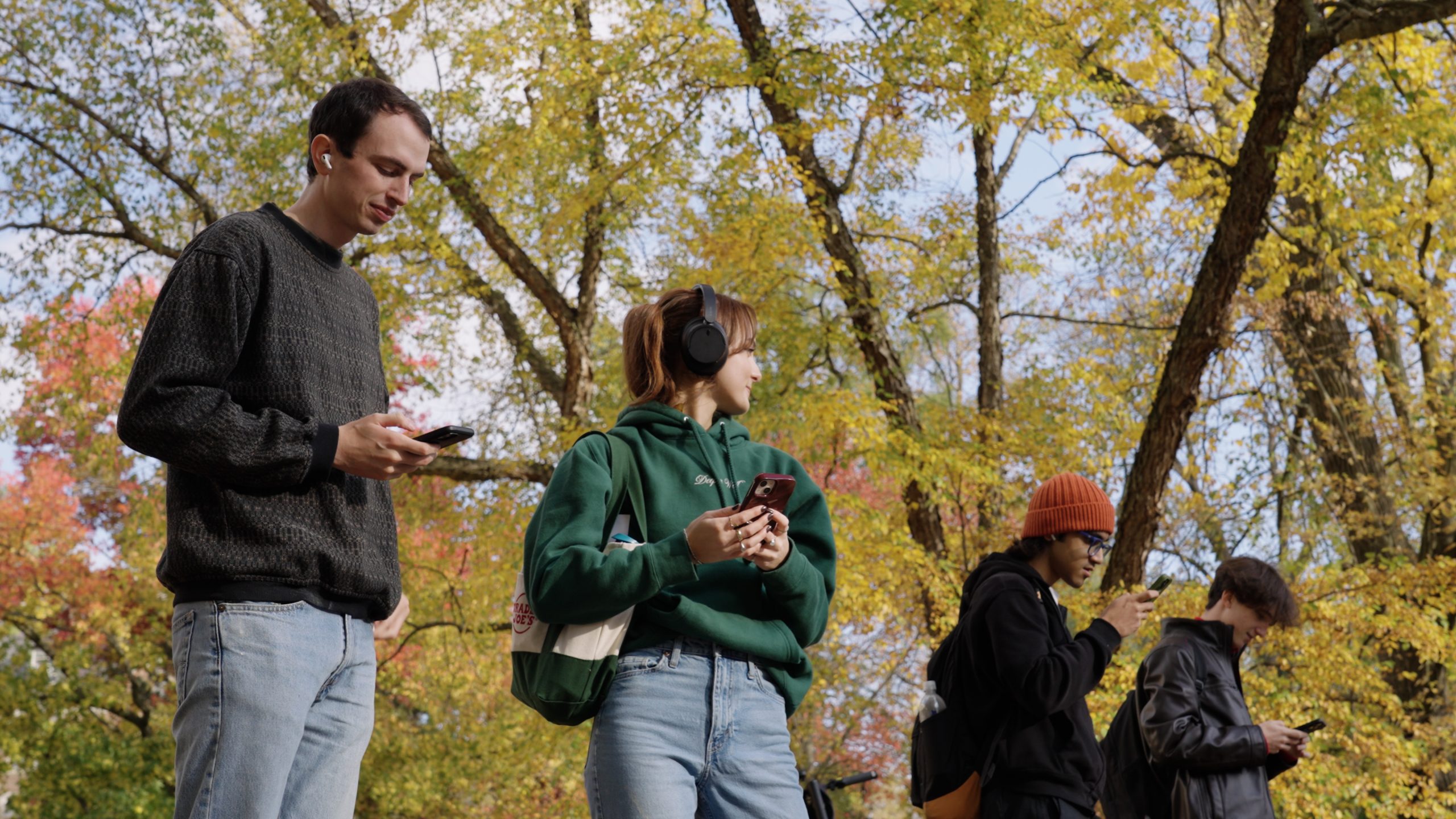 Four young adults are standing outdoors surrounded by trees with autumn foliage engaging with the Art Moves MSU app on their smartphones. A woman wearing a green hoodie has large black headphones on, while the other three men have their earbuds in.