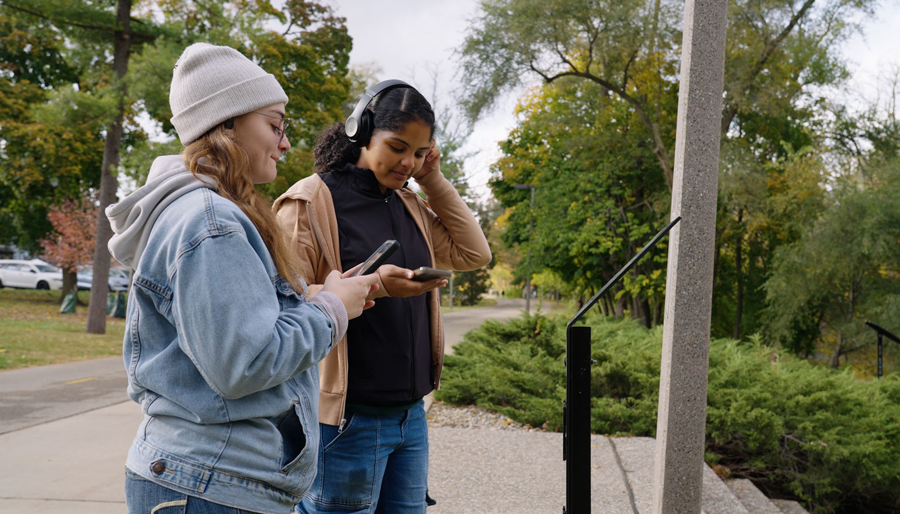 Two young women stand on the Red Cedar River Trail engaged with their smartphones. One woman, wearing a light blue denim jacket and a white beanie, looks at her phone with a smile. The other woman, in a brown jacket and black shirt, adjusts her headphones while focusing on her device.