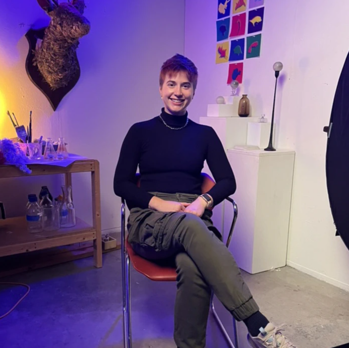 Hailey Becker sits in a brightly lit studio, wearing a black turtleneck and olive-green cargo pants, smiling at the camera. The studio features colorful artwork, shelves with glassware, and creative lighting.