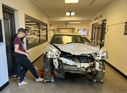 A heavily damaged car with a crumpled front end is displayed in a hallway. Two people stand nearby, observing the vehicle.