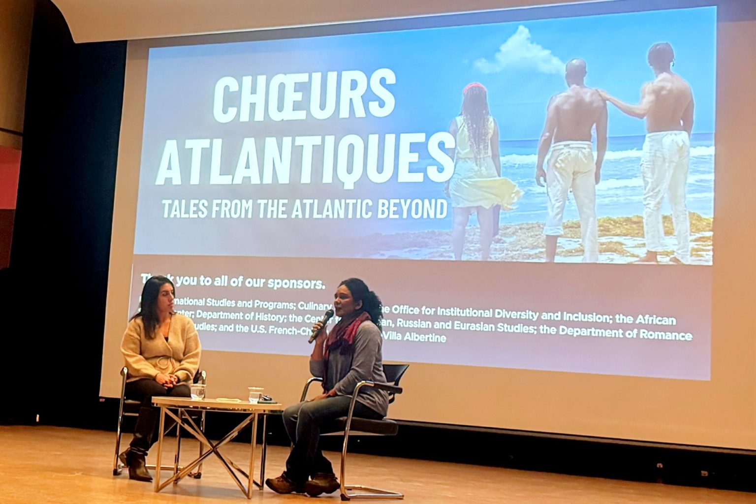 Two women sitting on a stage. One of the women is holding a microphone and speaking into it. Behind them is a large screen that has an image projected onto it with the words: 