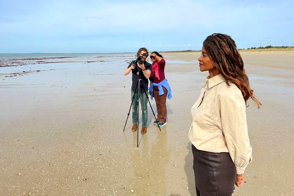 A woman with long braided hair stands on a sandy beach, gazing into the distance. Behind her a person films with a tripod-mounted camera while another crew member watches. The ocean and horizon are visible in the background.