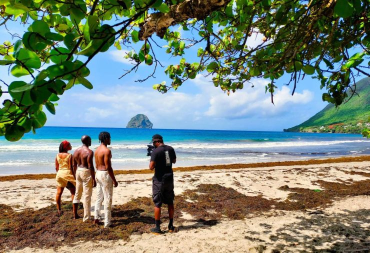 Three people, one woman and two men, stand on a beautiful beach with another man recording them. In the ocean there is a large rock in the center, with a green mountainside pictured to the right.