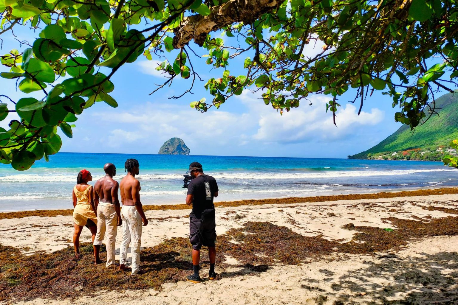 Three people, one woman and two men, stand on a beautiful beach with another man recording them. In the ocean there is a large rock in the center, with a green mountainside pictured to the right.