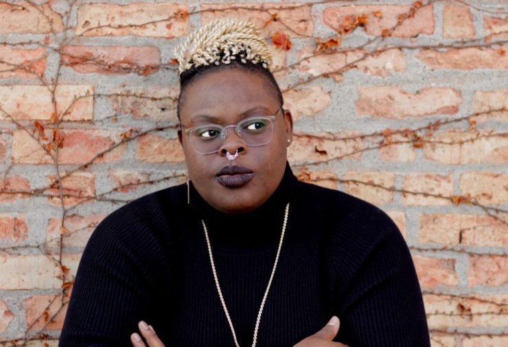 Head and shoulder photo of a Black woman wearing a black turtleneck, a long gold chain necklace, gold nose ring, glasses, and short hair on just the top of her head.