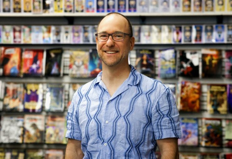 A man wearing glasses and a blue patterned shirt stands smiling in front of a wall of colorful comic books displayed on shelves.