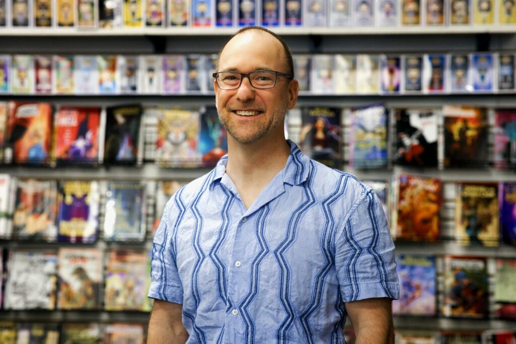 A man wearing glasses and a blue patterned shirt stands smiling in front of a wall of colorful comic books displayed on shelves.