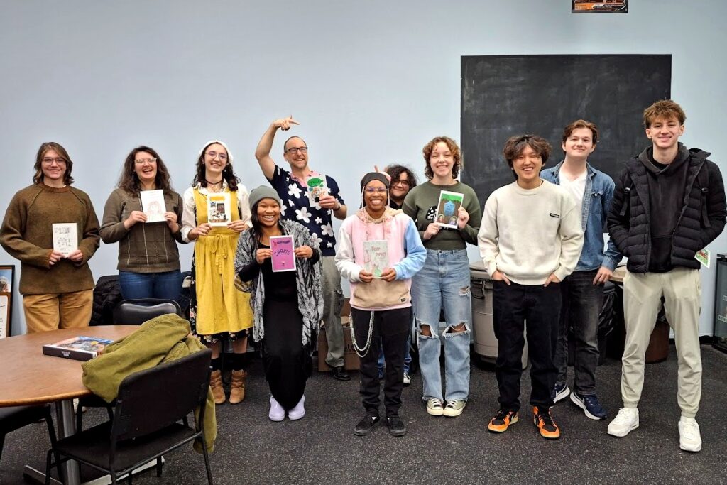 A group of students poses together in a classroom, holding comic books or with their hands in their pockets. In the center, a man, wearing a floral shirt, points with enthusiasm. A black board is visible in the background.