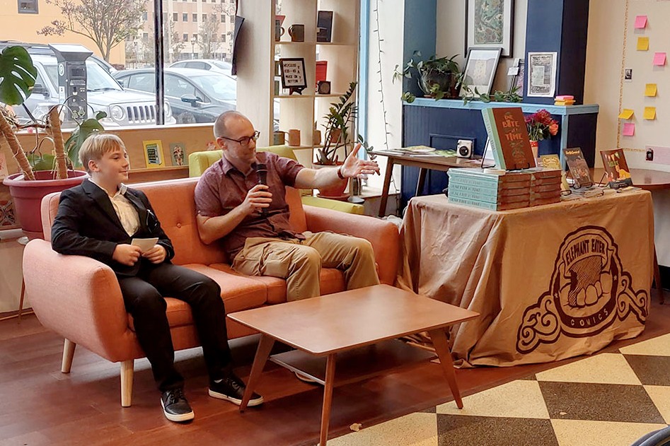 A man in a red shirt and khakis sits on an orange couch in a bookstore, holding a microphone and gesturing as he speaks. Next to him, a young man in a suit listens. A table nearby displays copies of One Bite at a Time with an Elephant Eater Comics-branded tablecloth.