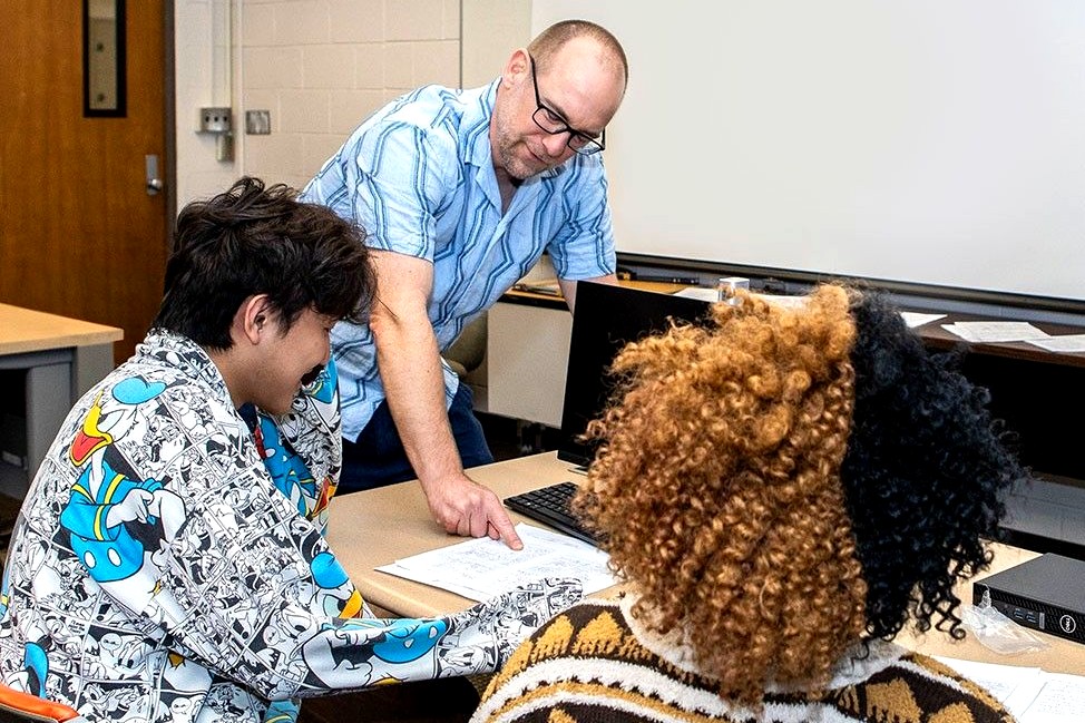 A man wearing glasses and a blue patterned shirt, leans over a desk while engaging with two students. One wears a comic-themed hoodie, and the other has curly hair. Papers and drawings are spread out in front of them.