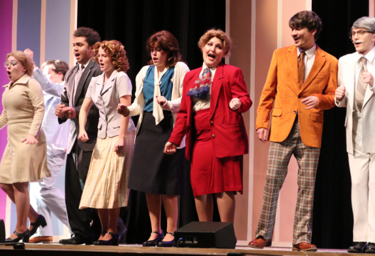 Seven people singing and dancing on stage. They are dressed in 1980's office attire.