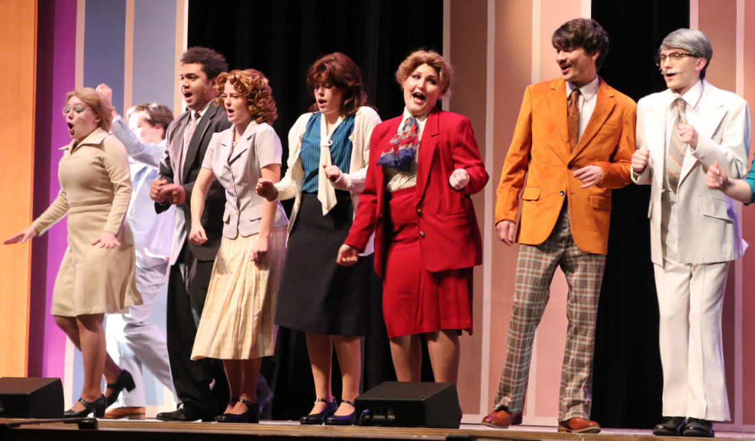 Seven people singing and dancing on stage. They are dressed in 1980's office attire.