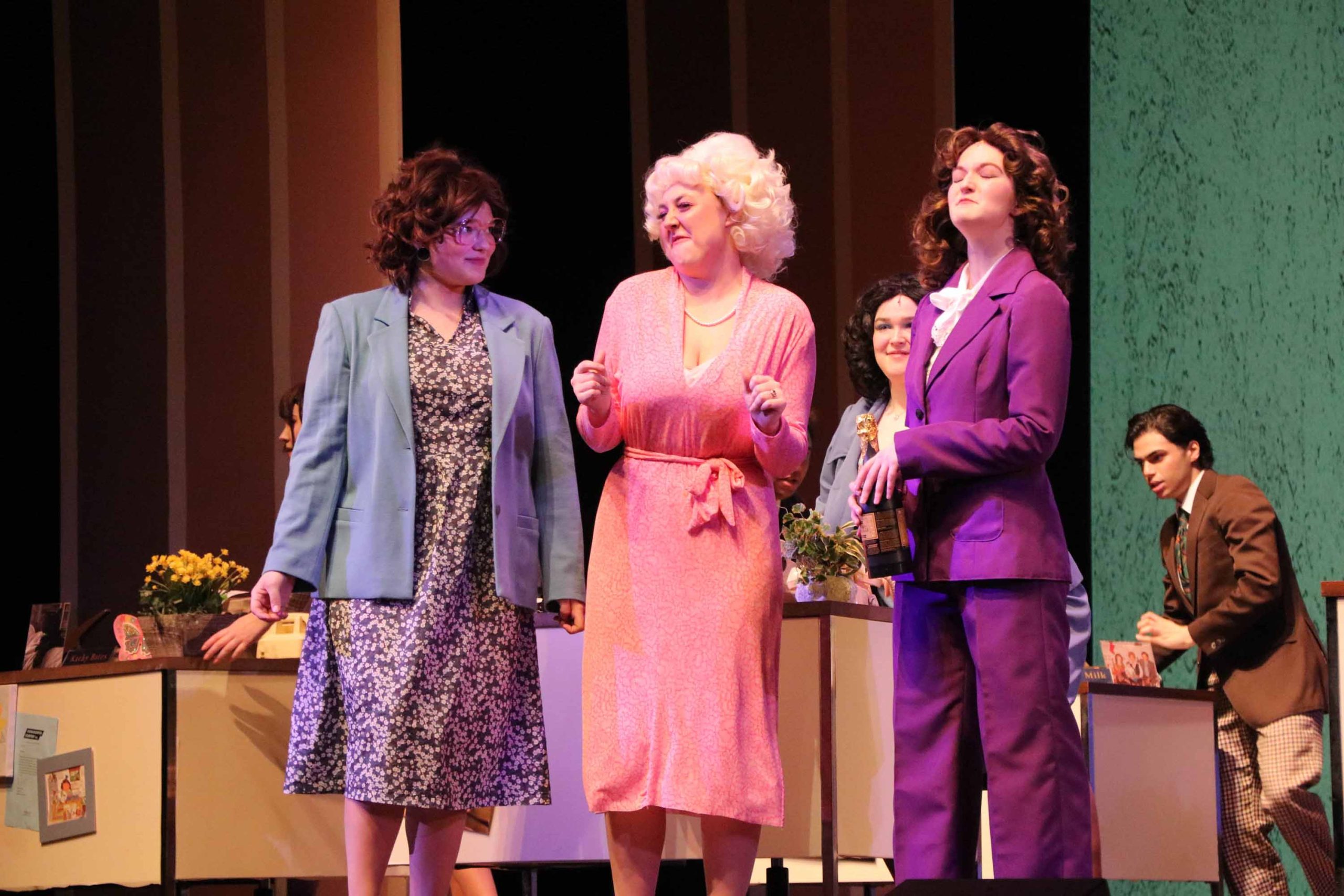 Three women stand on a theatre stage. From left to right, the first woman is wearing a long black and white speckled dree with a blue blazer. The second is wearing a pink dress with a bow tied at the waist. The last woman is wearing a purple pant suit.