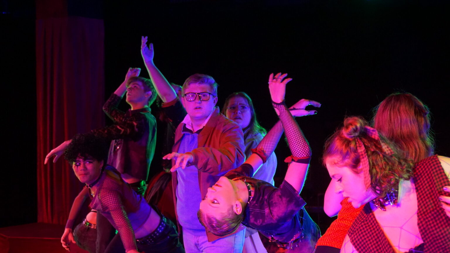 A group of performers, dressed in edgy, punk-inspired costumes, strike expressive poses on stage. The lighting casts a dramatic glow in red and purple tones, enhancing the theatrical atmosphere.
