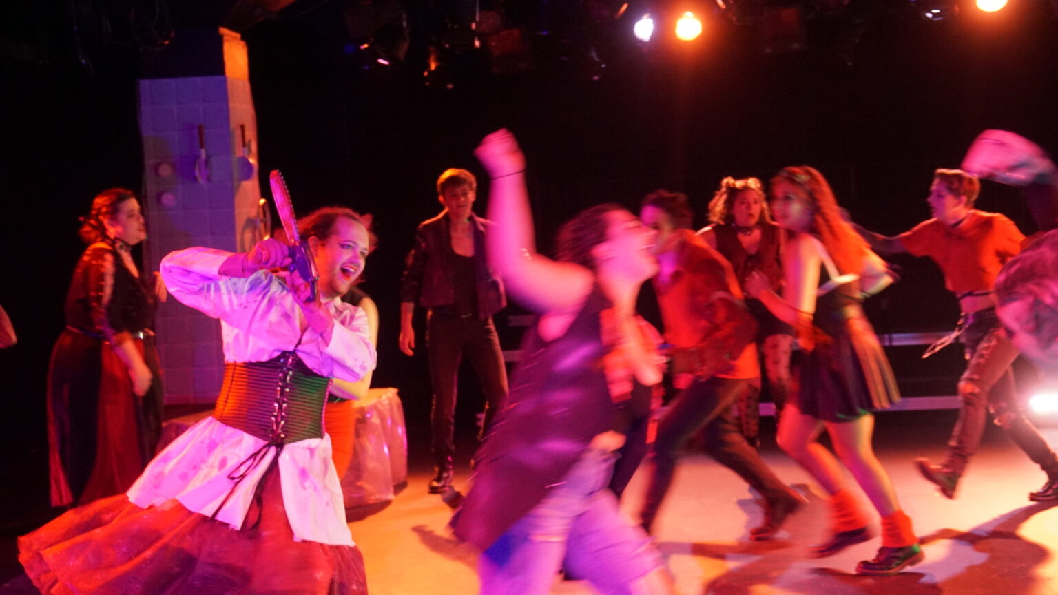 An intense stage scene shows an an actor wielding a large prop weapon as a group of performers around her move in dynamic, blurred motion. The lighting is warm, with orange and red hues creating a chaotic energy.