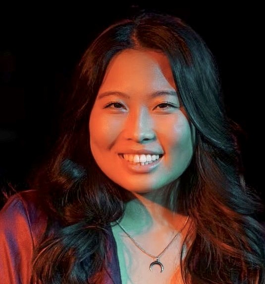 A portrait of a woman smiling against a dark background, lit with dramatic lighting in teal and orange. She wears a deep red jacket, black shirt, and crescent moon necklace, with soft waves in her long dark hair.