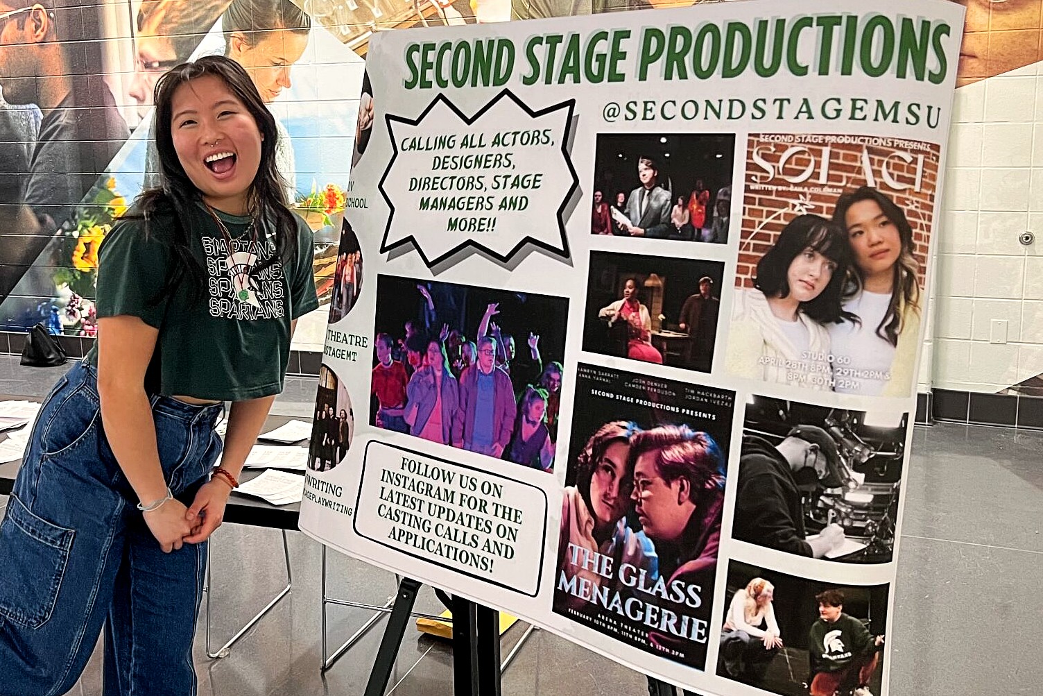 A woman wearing a green shirt with blue jeans stands smiling in front of a poster board titled 