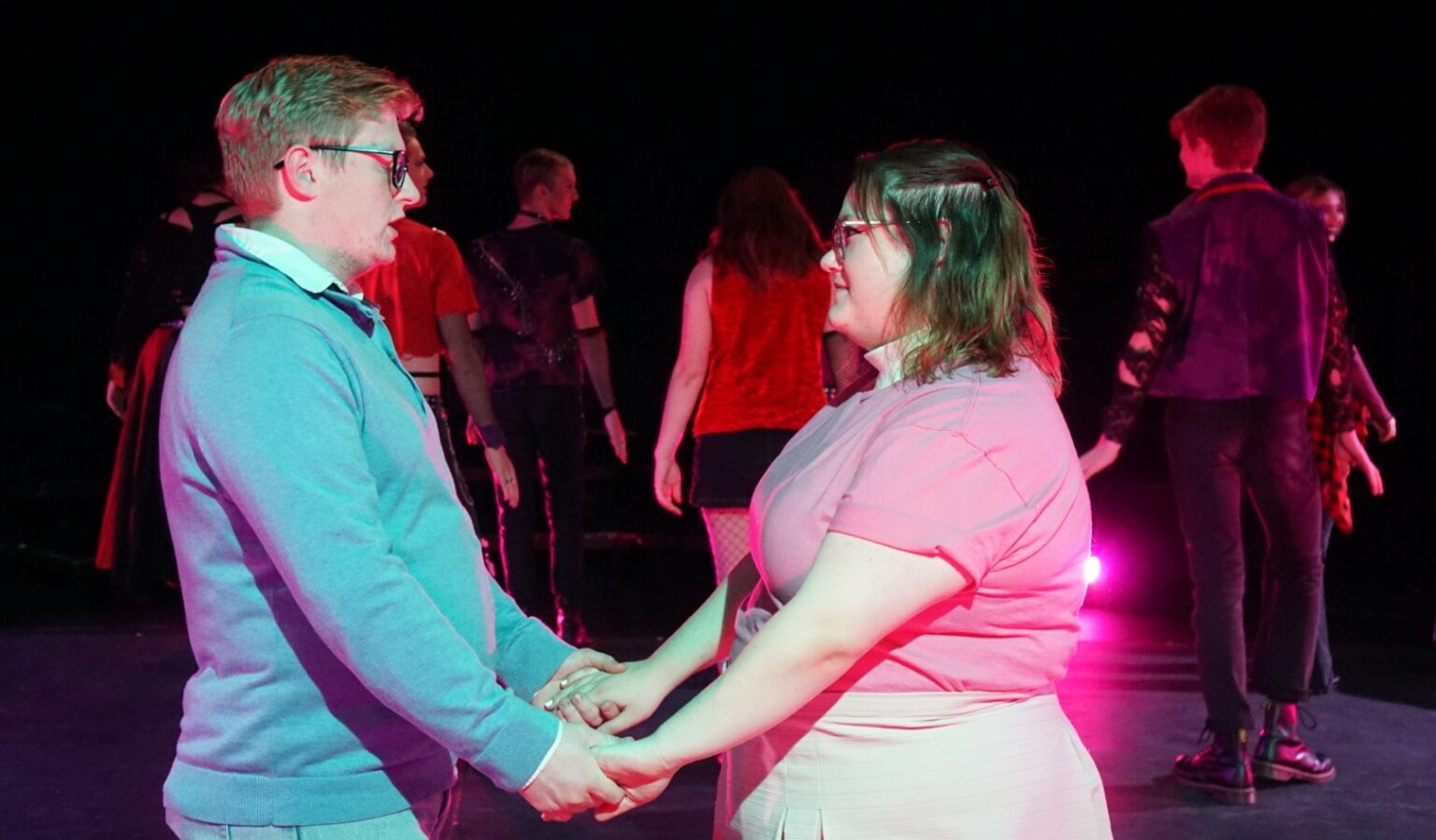 Two actors, a man in a blue sweater and a woman in a pink top, hold hands and gaze at each other center stage. Behind them, several performers in red and black costumes move under pink stage lighting.