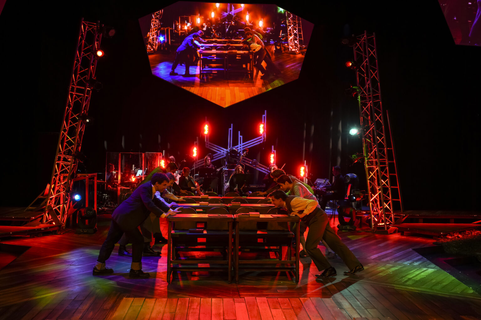 On a stage with red and green lighting, actors in formal clothing push desks together. Above them, a mirrored projection of the same scene is displayed on a suspended screen.