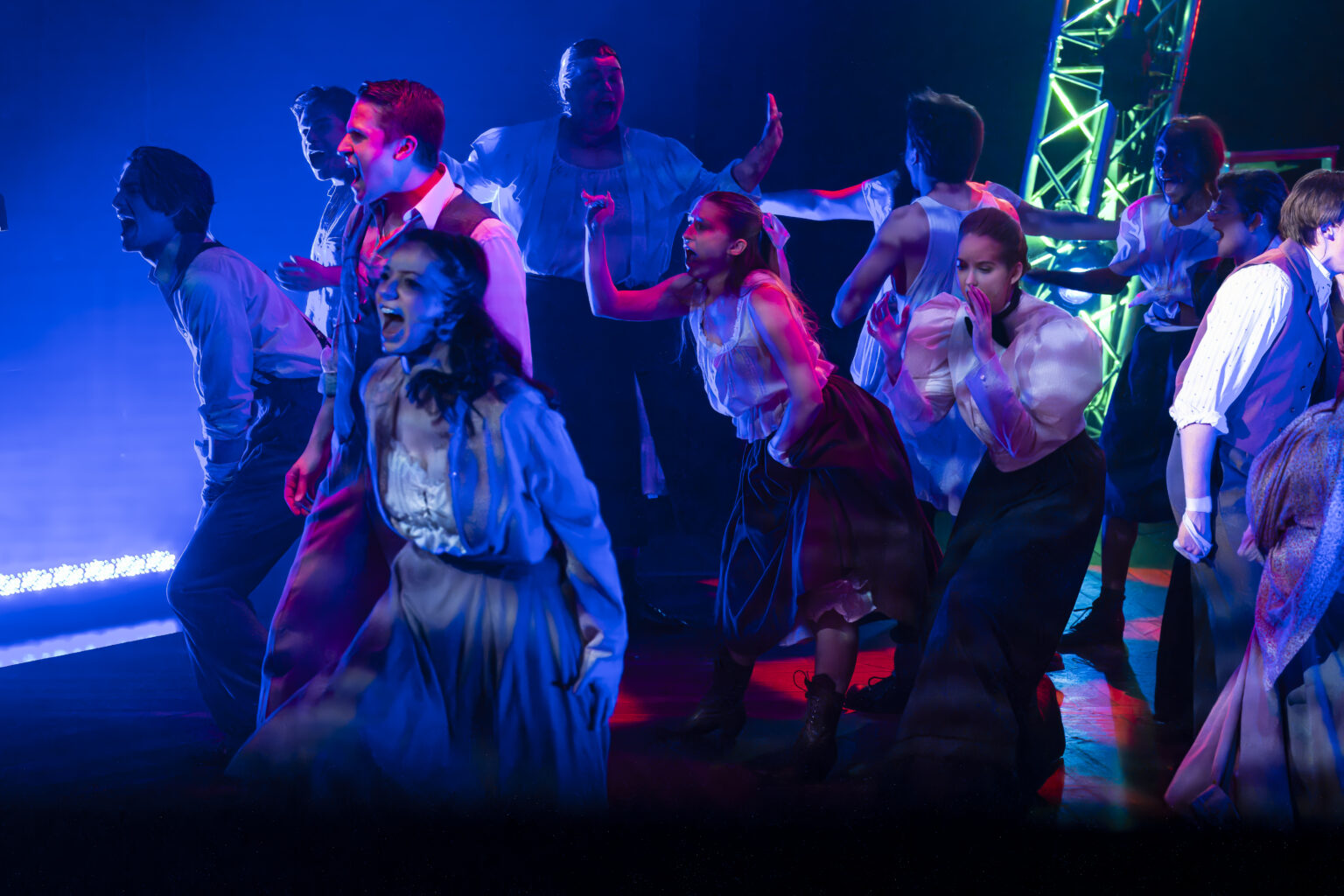 A group of actors dressed in period-style clothing perform a scene under vibrant blue and magenta lighting.