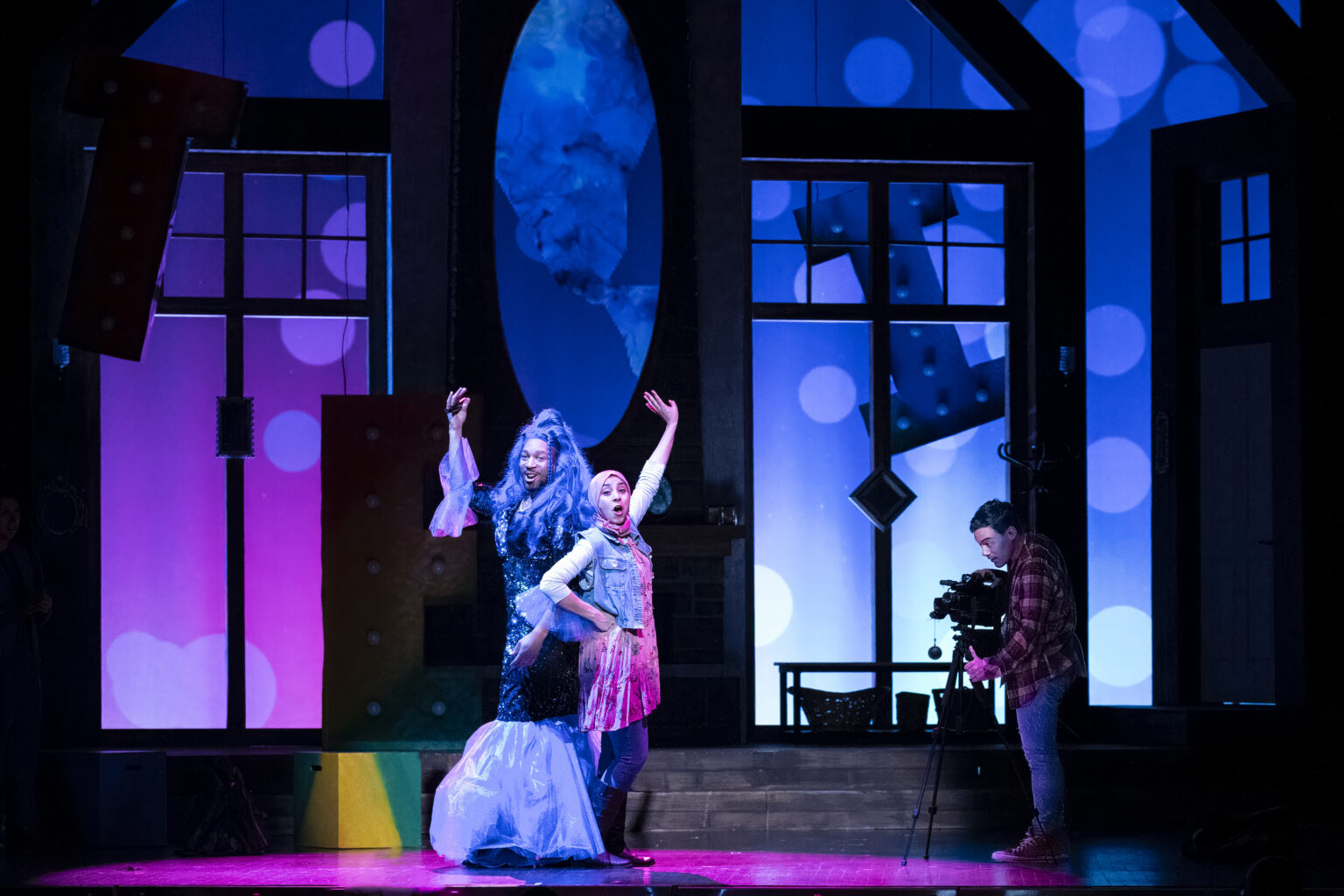 A performer in a glamorous gown and a long blue wig poses next to a performer wearing a denim vest and tie-dye skirt. A cameraman in a flannel shirt films the scene, set against a blue and purple backdrop.