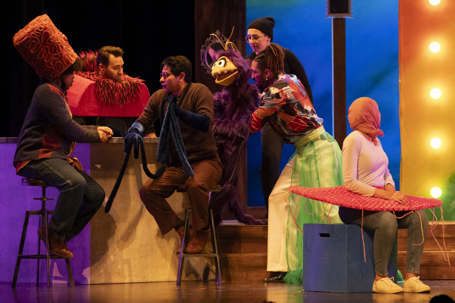A whimsical puppet scene on stage features actors interacting with colorful, oversized puppets.