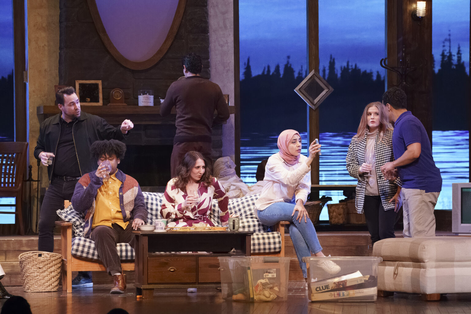 Actors perform a living scene in a cozy, rustic set. The backdrop shows a painted lake view through tall windows.