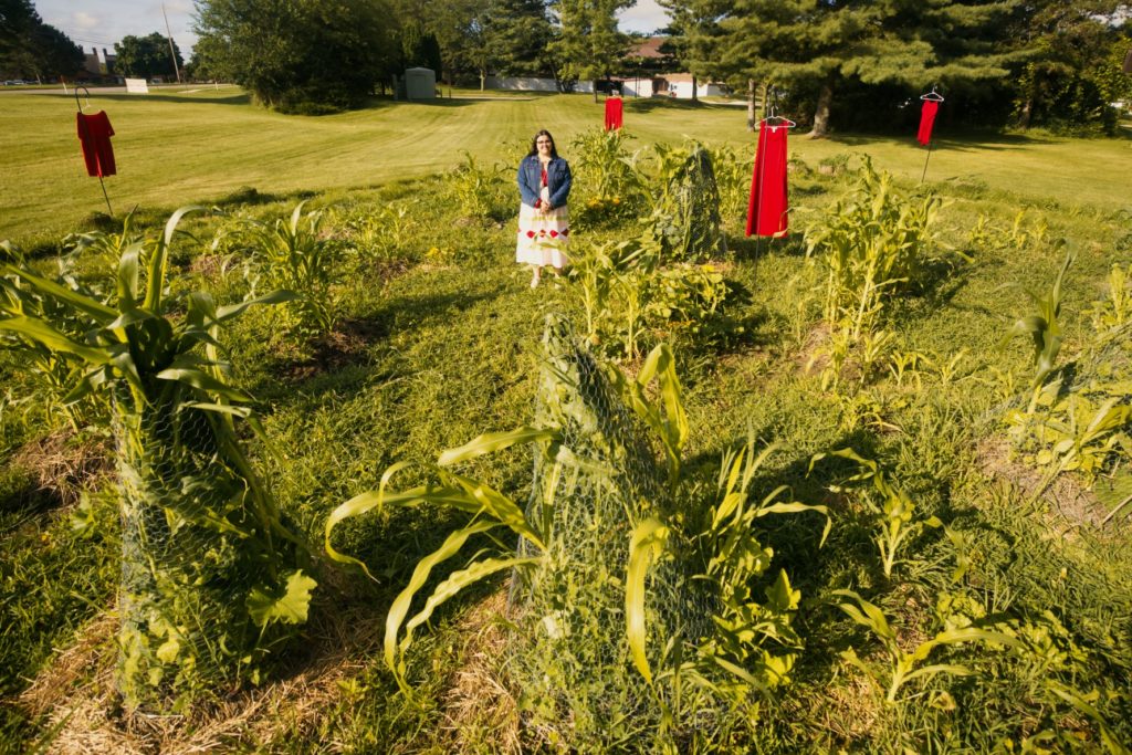 A woman stands in a garden surrounded by green stalks of corn. There are four red dresses hanging in the background.