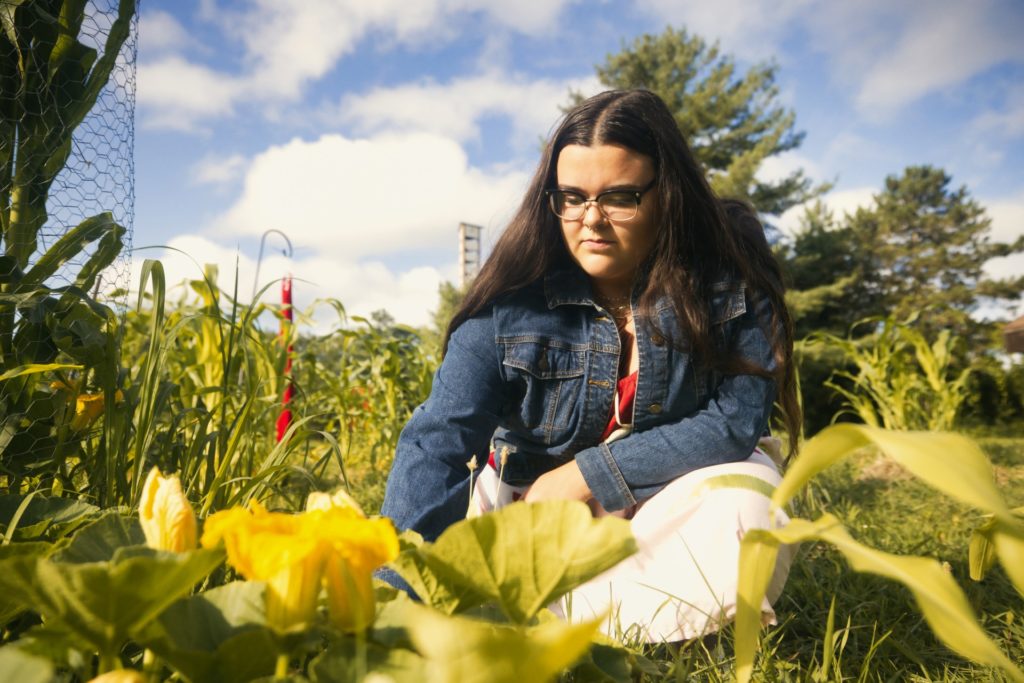 A woman crouched down in a garden in front of squash vines and flowers.