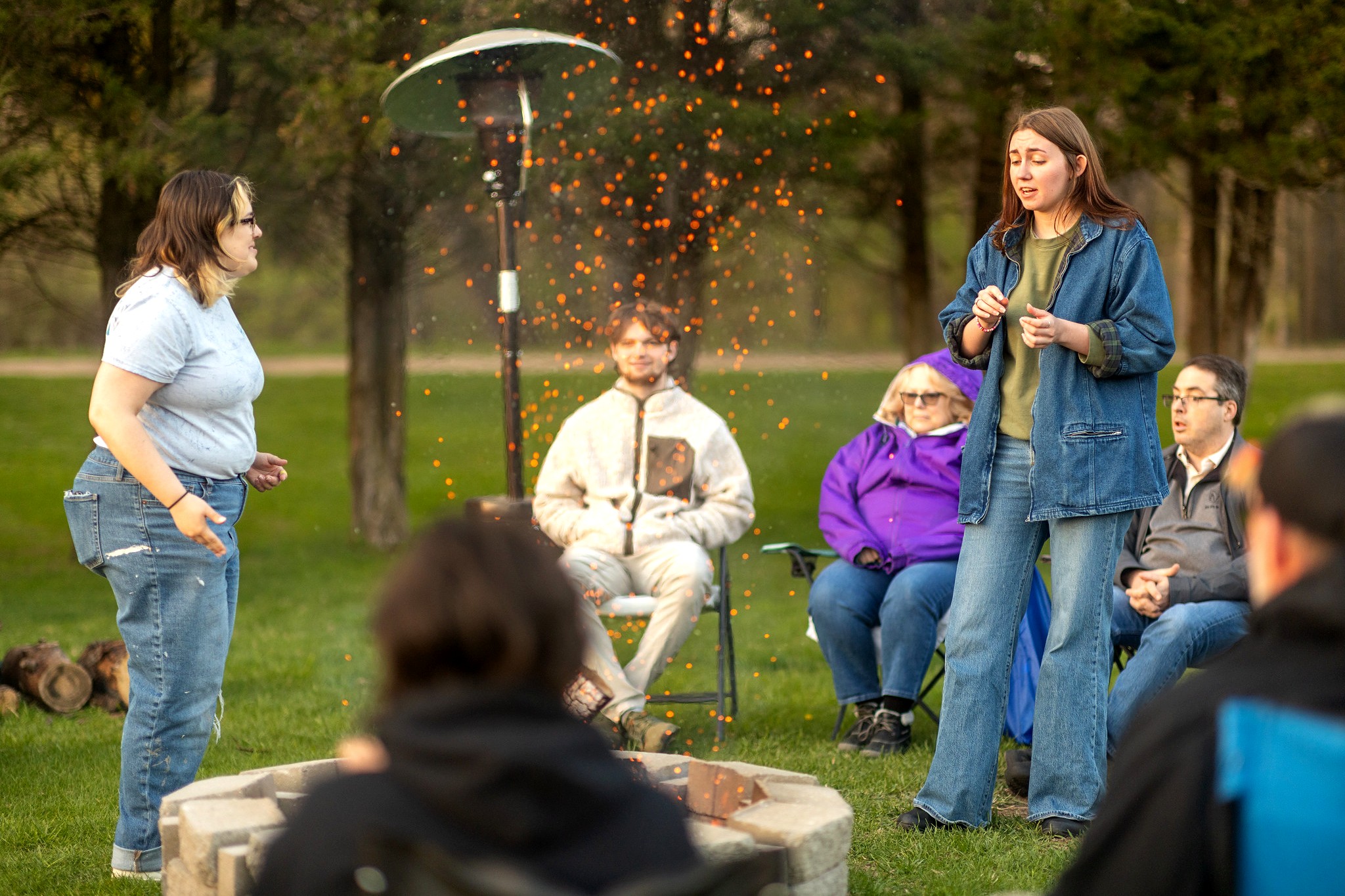 Two individuals stand and perform near a fire pit outdoors, with orange embers rising in the air. They are surrounded by seated spectators in lawn chairs, dressed for cool weather under trees and portable heaters.