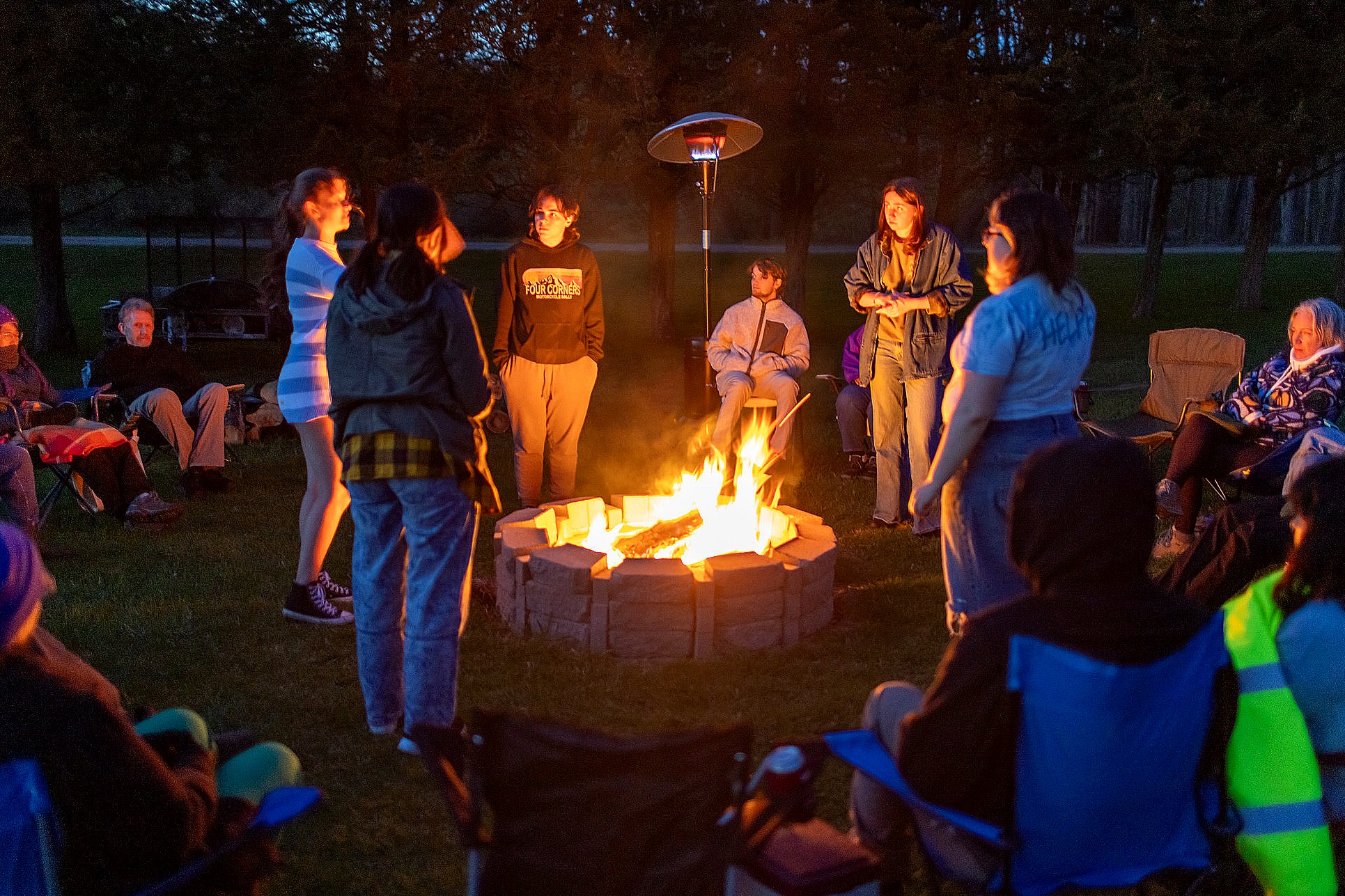 A group of people stand around a fire pit at night, warmly lit by the flames. Others sit in a circle of camping chairs attentively watching. The gathering takes place outdoors on a grassy field, surrounded by trees and illuminated by a tall outdoor heater.
