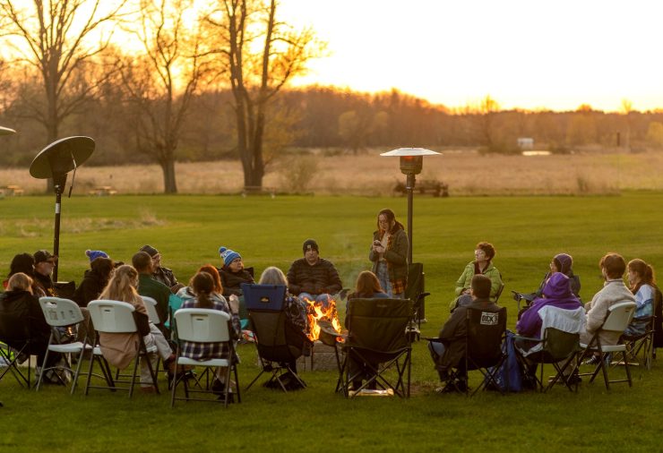 A large circle of people sits in folding chairs around a fire pit on a grassy field at sunset. One person stands and speaks while others listen, with trees and open landscape stretching into the distance.