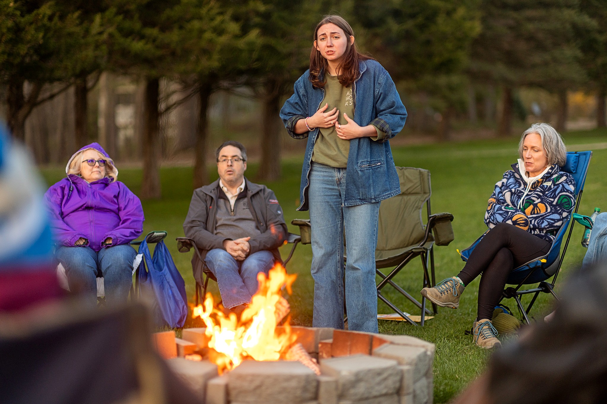 A person stands and speaks near a fire pit, using hand gestures, while others seated nearby listen attentively. The group is gathered in lawn chairs on a grassy field with trees in the background, dressed warmly for the evening.