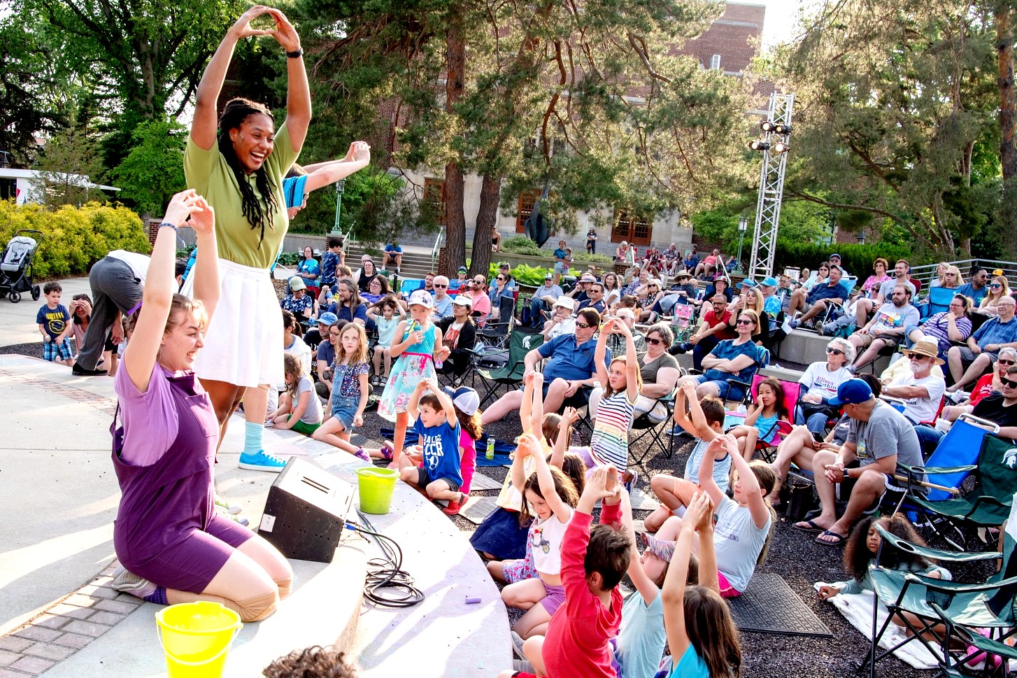 A lively outdoor theatre scene where two performers lead a group of children in interactive movements. The crowd includes families and children, many of whom are mimicking the performers' arm movements. The setting is sunny, with trees, a campus building, and stage lighting the background.